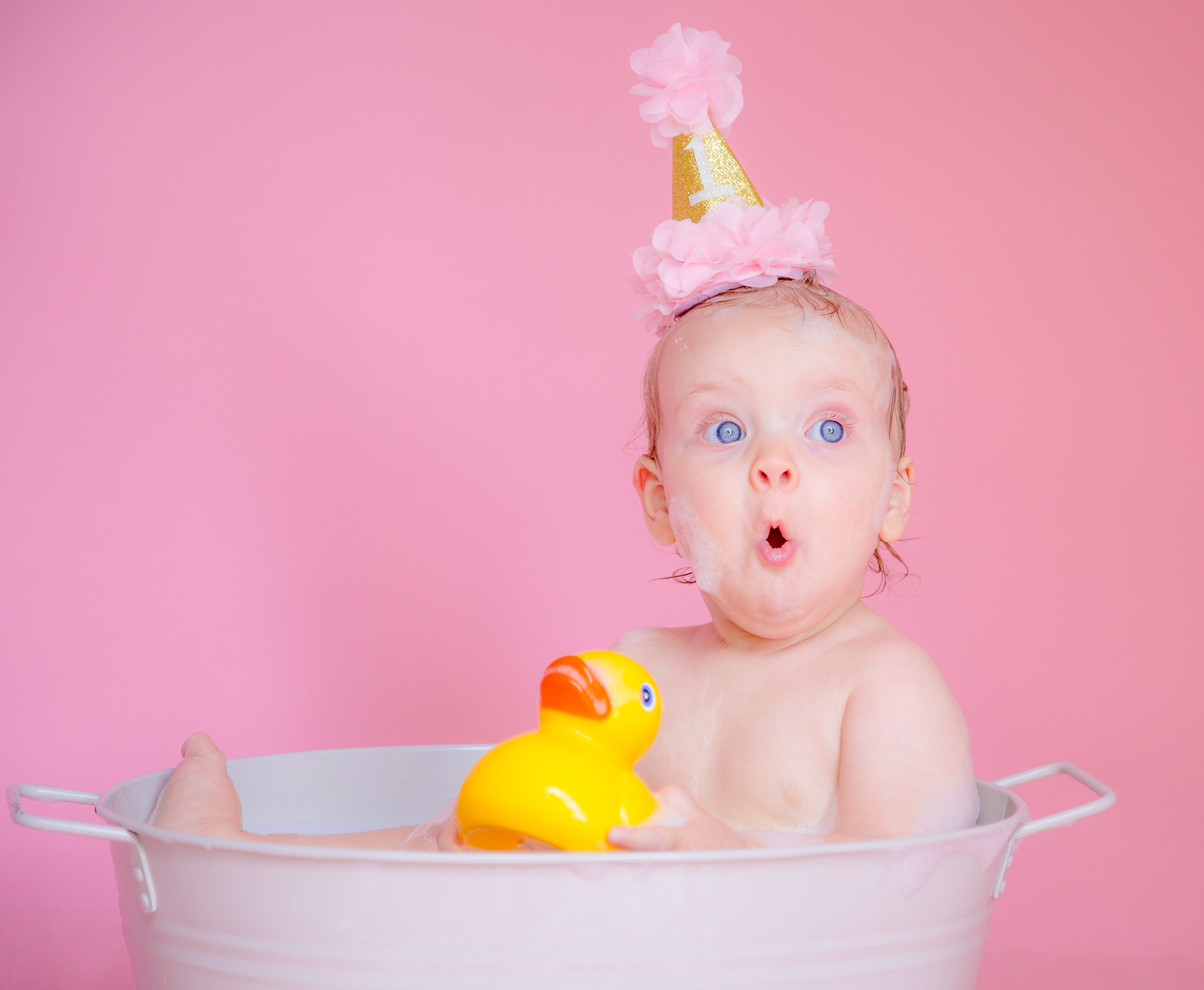 A baby girl sits in the bath with a first birthday hat on for her sevenoaks cake smash photos