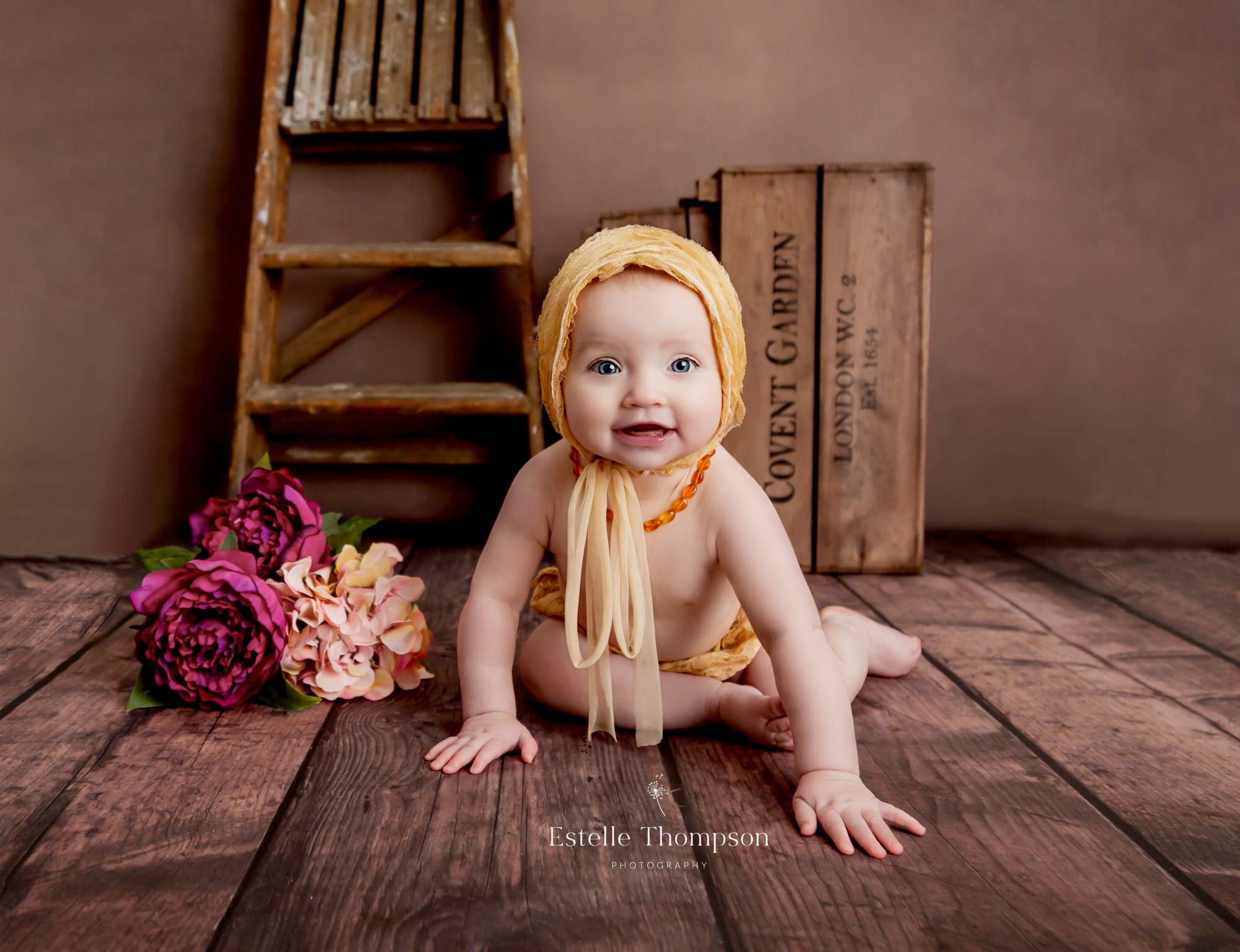 Baby girl wearing a gold bonnet crawls on the floor smiling by Sevenoaks baby photographer