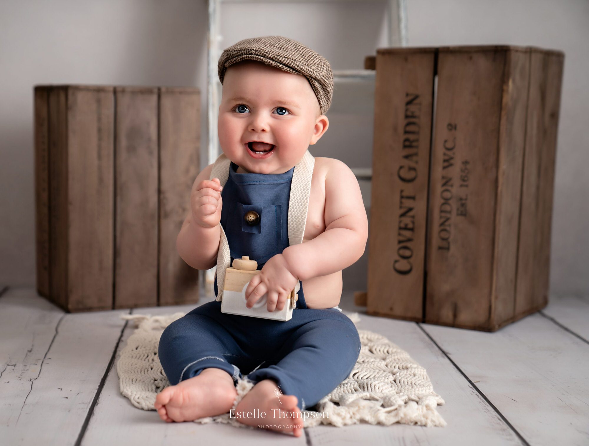 Baby boy sits on the floor laughing holding a wooden camera toy by Sevenoaks baby photographer