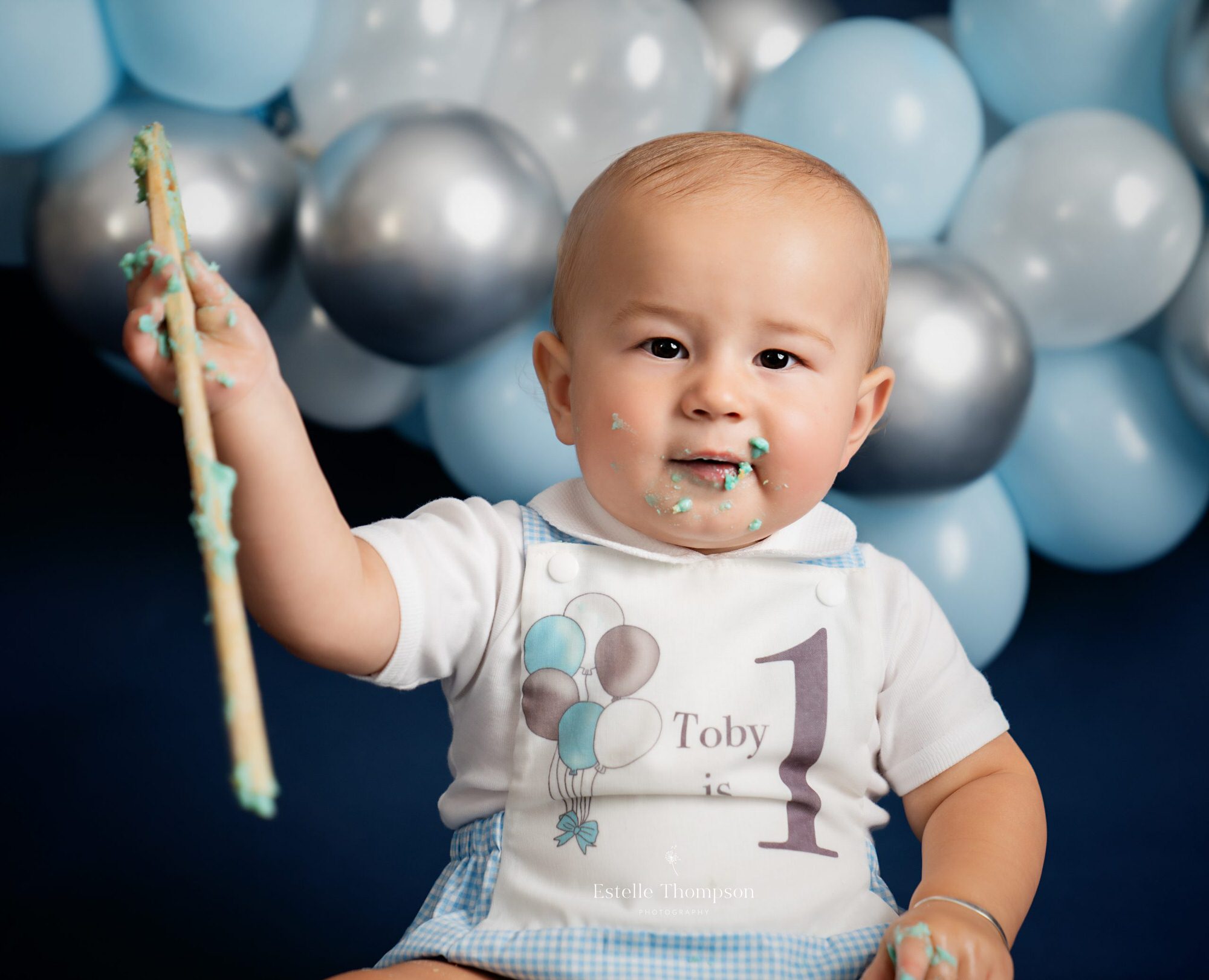 Baby boy holds up a wooden spoon he is using to smash a cake in his cake smash in sevenoaks