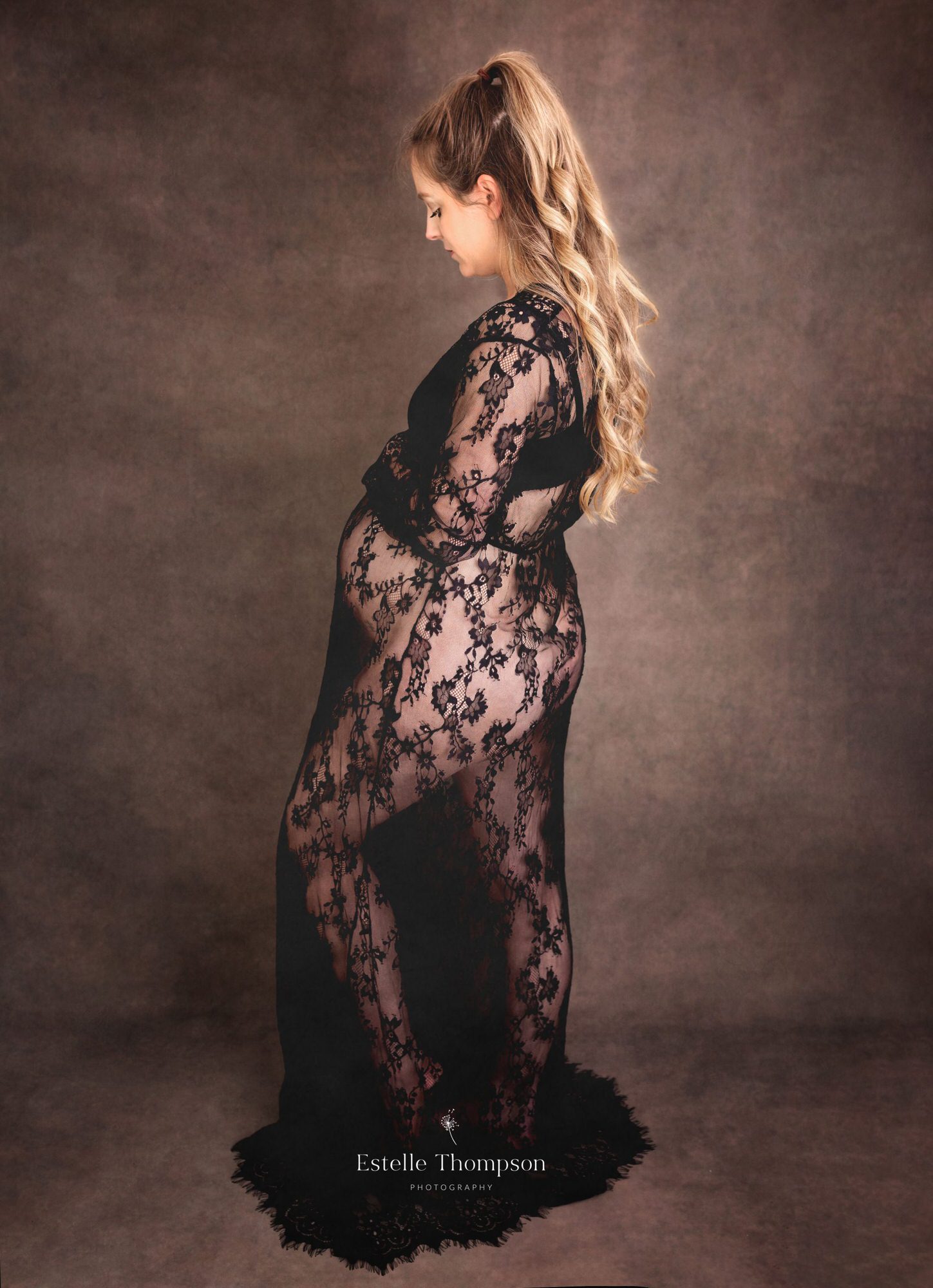 A pregnant lady wearing a black lace dress holds her bump in a Kent maternity photography studio