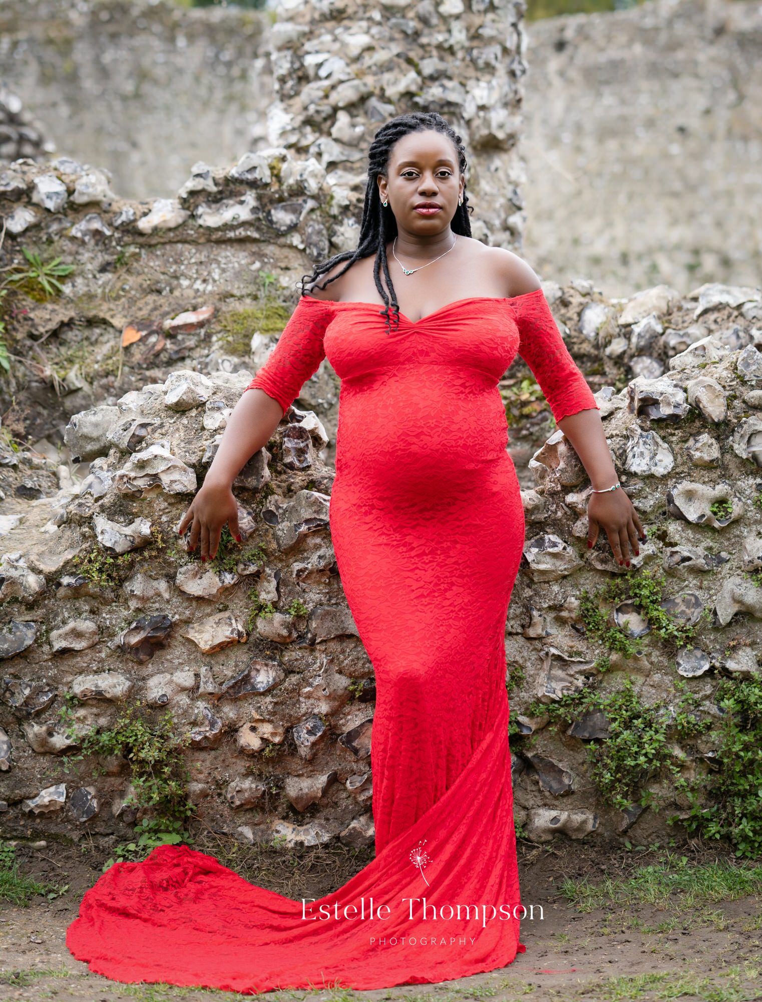 A pregnant lady wears a red lace dress and leans against a wall having her maternity photos taken by Sevenoaks maternity photographer