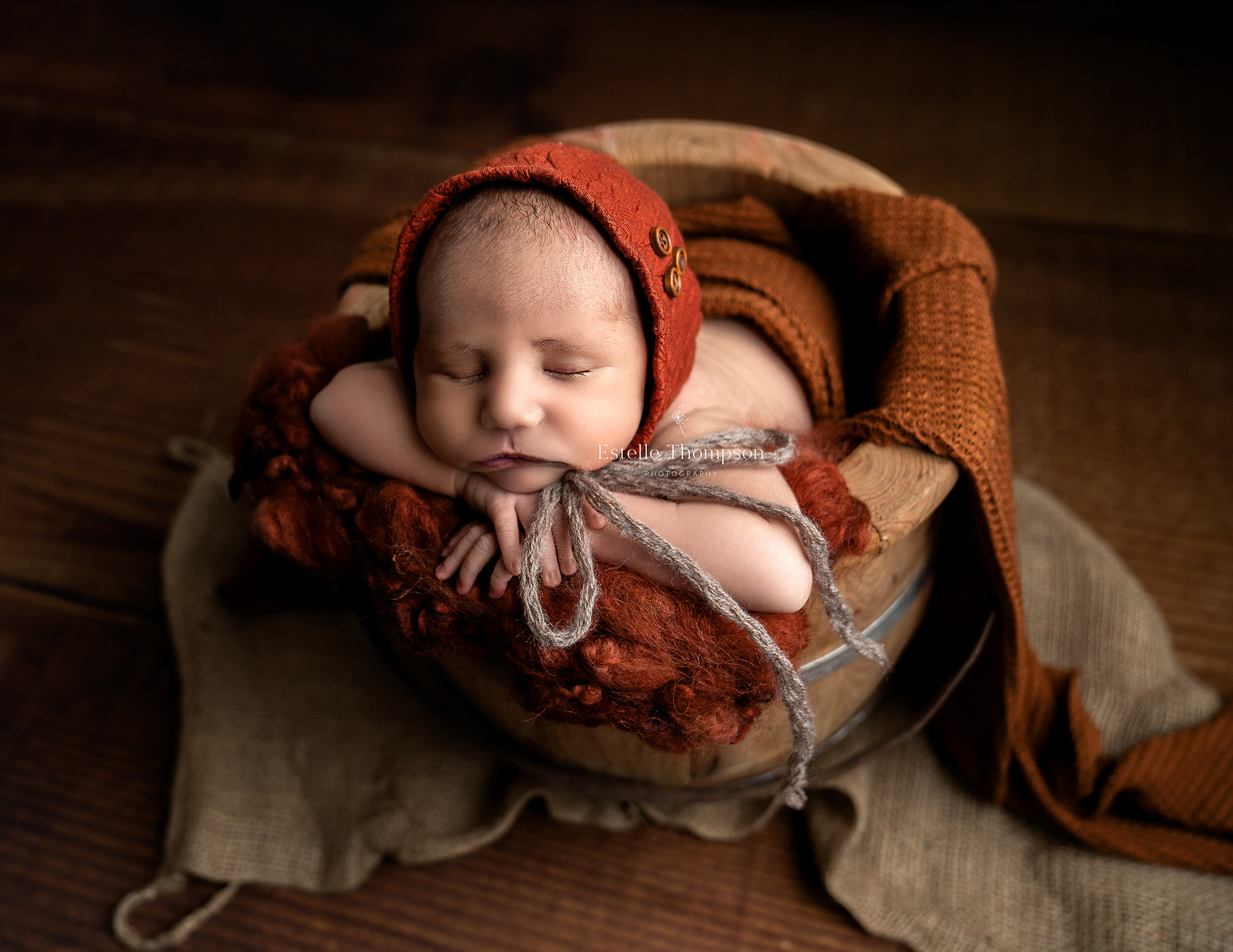 Baby boy wearing a rust coloured bonnet laying in a wooden bucket